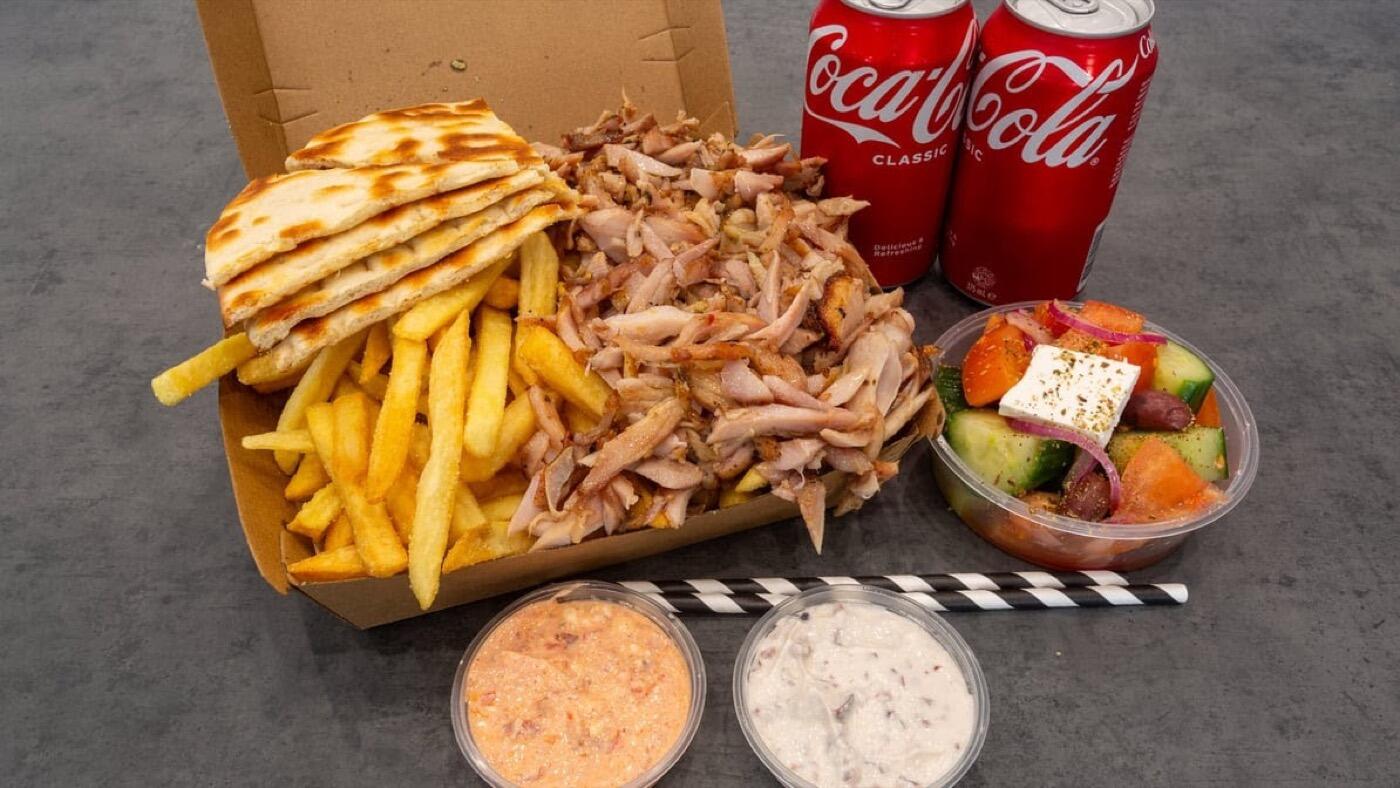 A meal pack: a box of chips, flatbread and sliced meat, two cans of Coke, a small Greek salad with feta and two dips