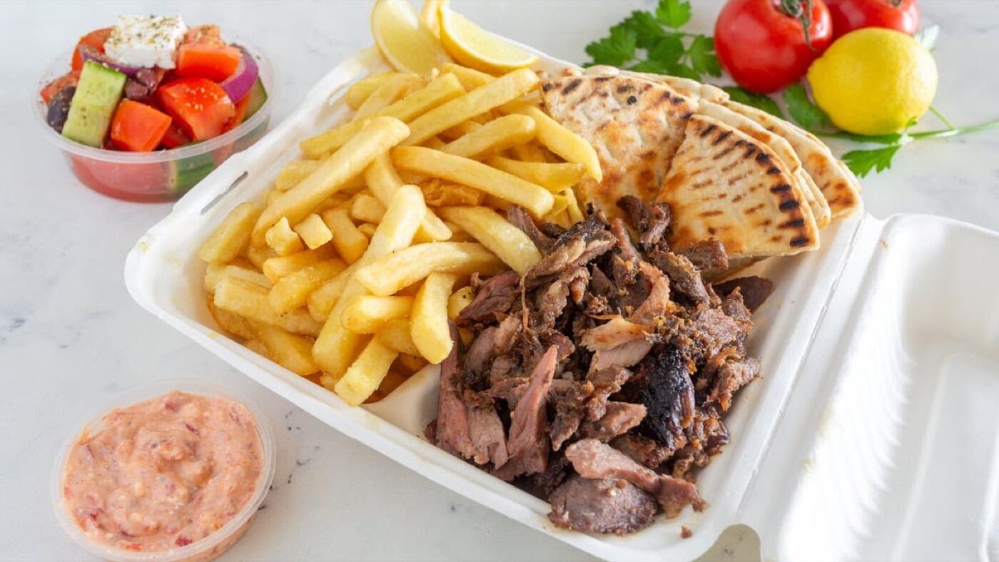 A takeaway box of chips, grilled pita bread and sliced meat, with a pot of pink dip, a small salad and a lemon
