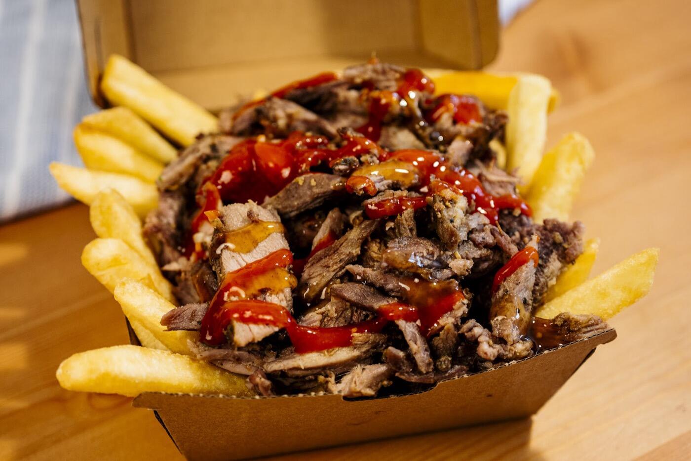 A cardboard box of sliced grilled meat with red and yellow sauces over chips