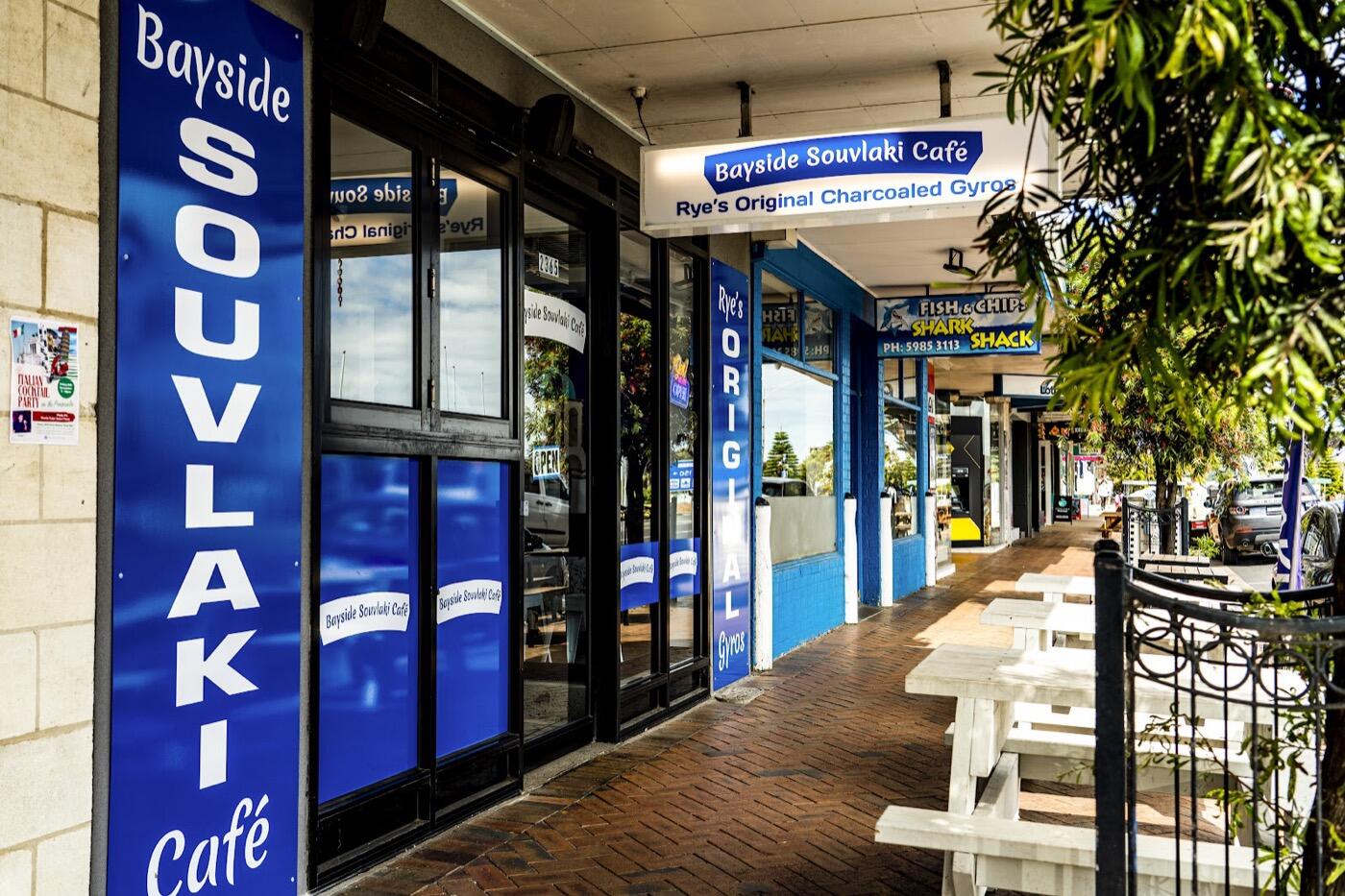 The shopfront with a Bayside Souvlaki Cafe sign above big glass windows, tall blue banners and white outdoor benches