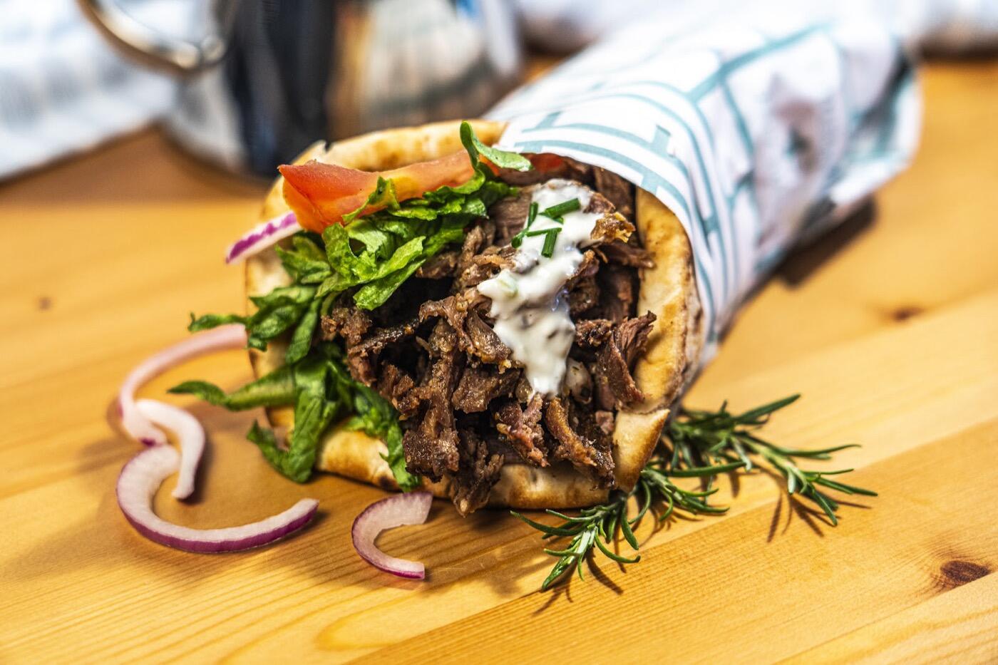 A close-up of a souvlaki wrap filled with grilled meat, lettuce, tomato, red onion and white sauce, with a sprig of rosemary