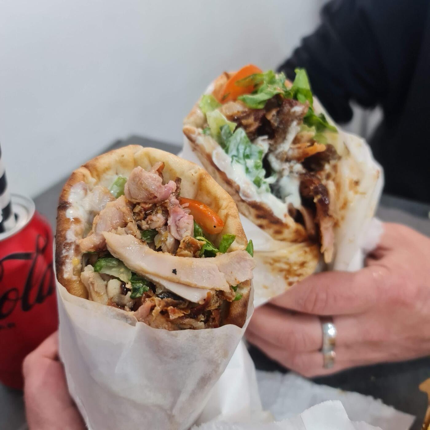 Two souvlaki wraps in paper held in a hand, one with sliced meat and salad and the other with meat and a creamy sauce, with a red can beside them
